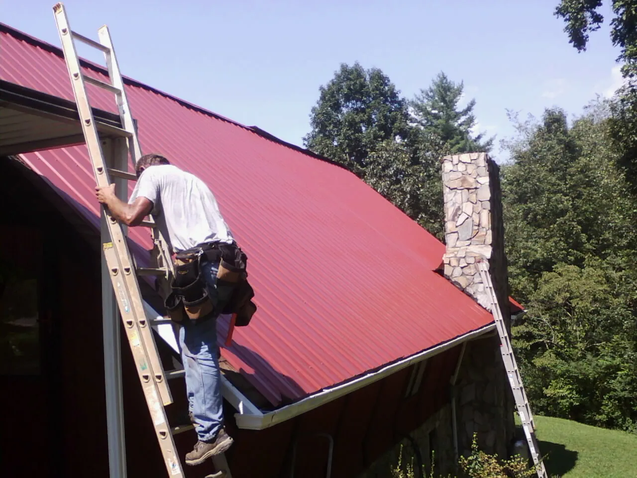 Roofer on a ladder beside a red metal roof and stone chimney — Soffit & Fascia Repair by Roofing Brothers in Sun Valley